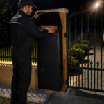 Modern secure parcel delivery box installed beside a black metal gate at a residential property in London, with a courier placing a parcel inside during the evening.