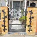 Black decorative wrought iron pedestrian gate with floral leaf details mounted between yellow brick pillars at the entrance of a luxury London home.